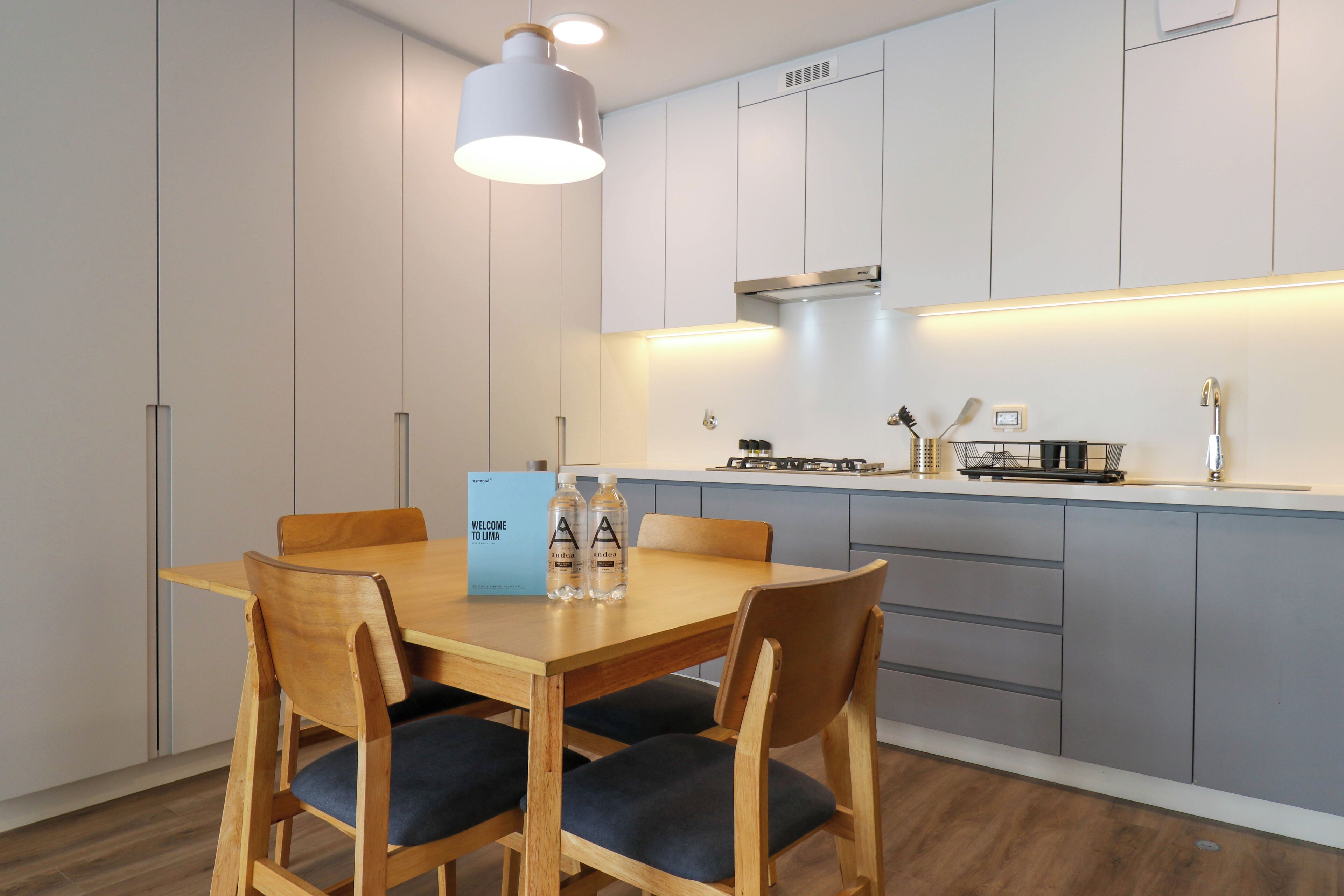 A modern kitchen with a wooden dining table and chairs, a hanging light fixture, and a welcome sign.