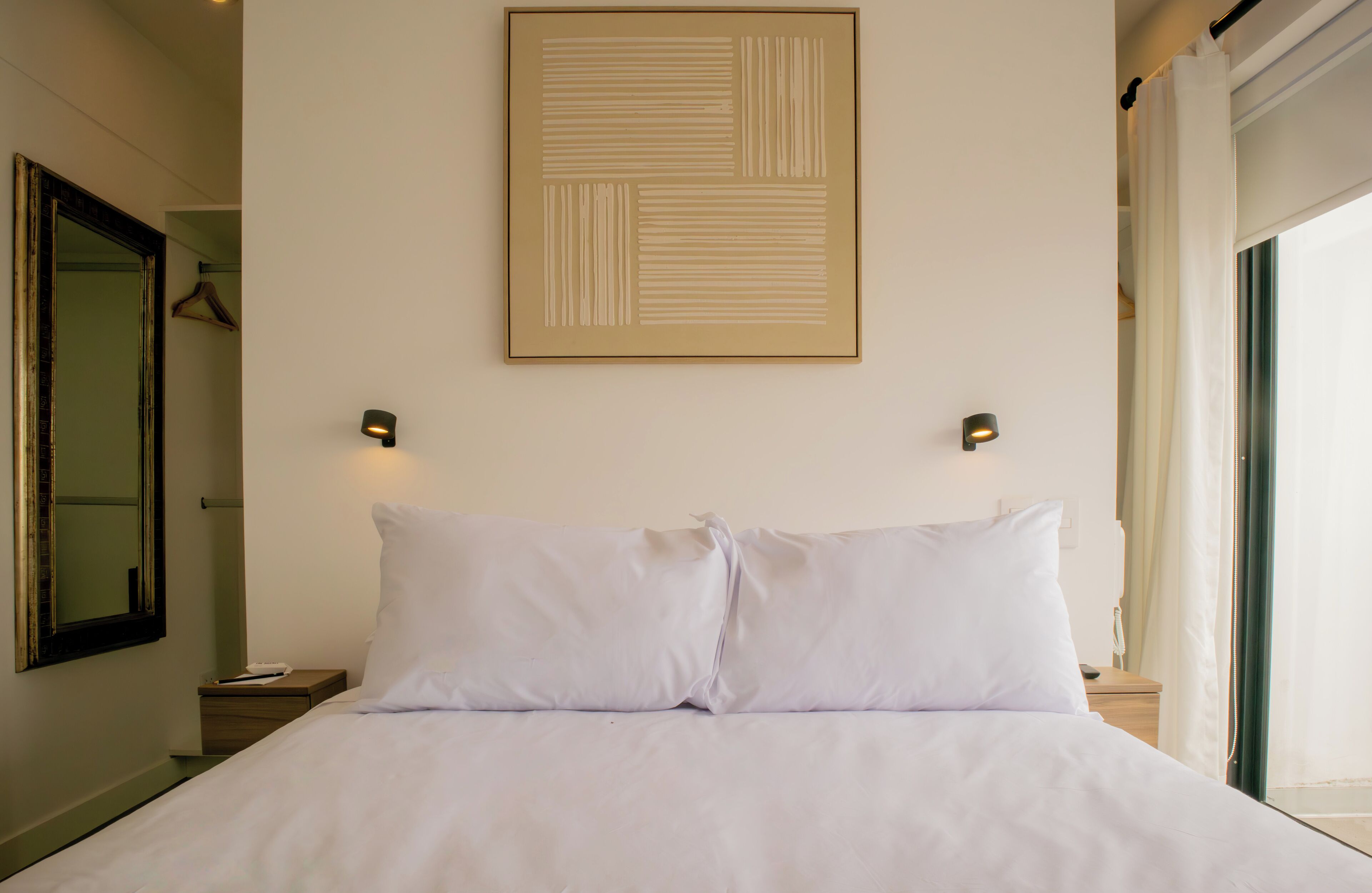 A neatly made bed in a hotel room with a large framed artwork above it and a mirror on the left wall.