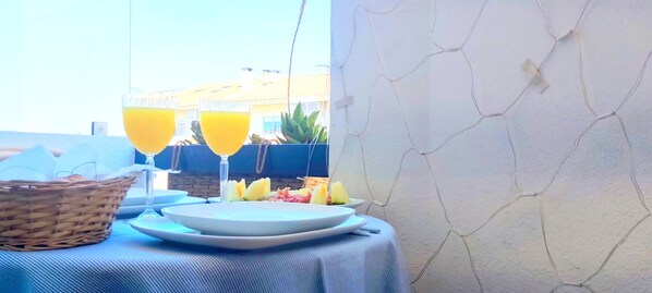 A table set with plates, a basket, and glasses of orange juice, with a view of buildings and plants in the background.