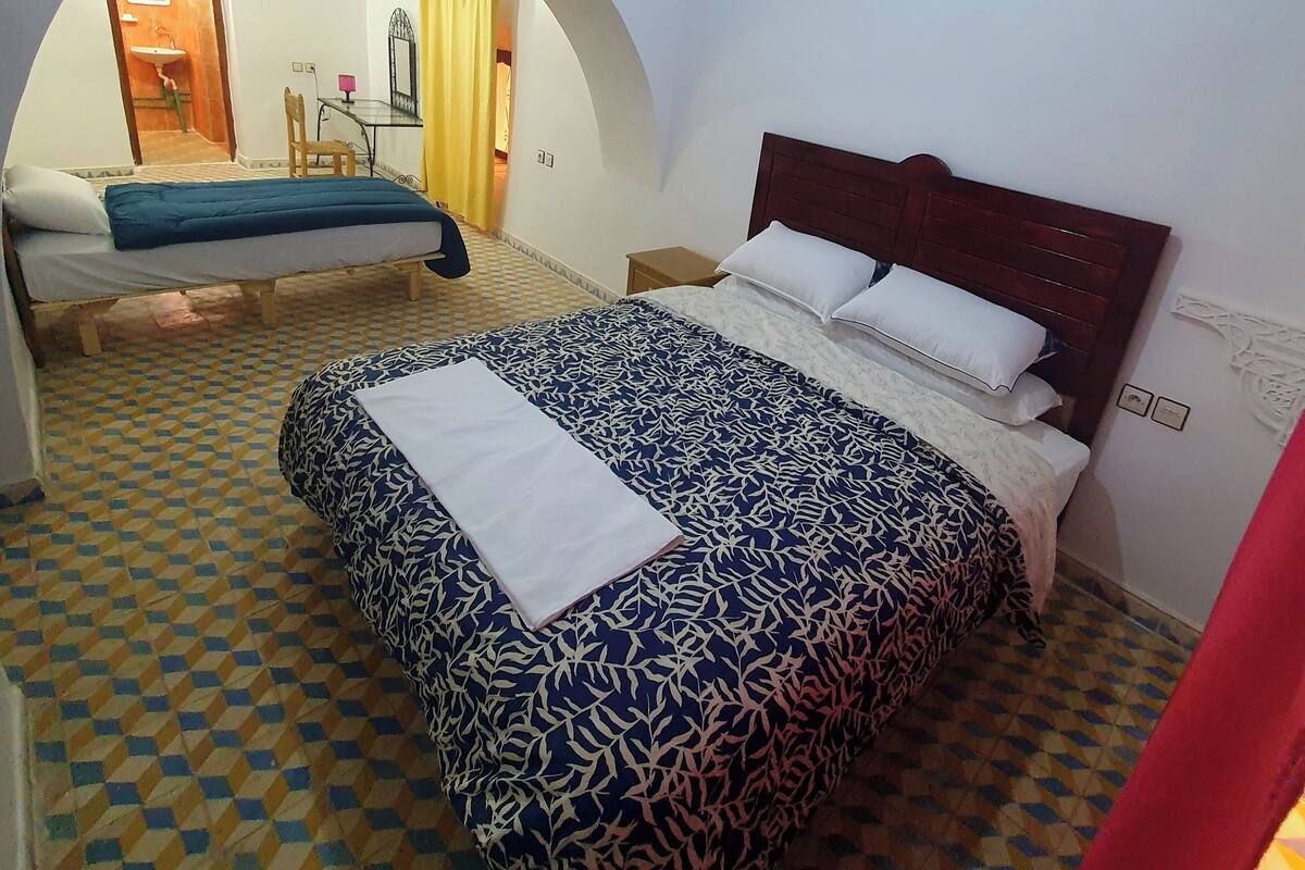 Ein Hotelzimmer mit einem großen Bett, einem Einzelbett, einem Schreibtisch mit Stuhl und einem durch einen Bogen erreichbaren Badezimmer.