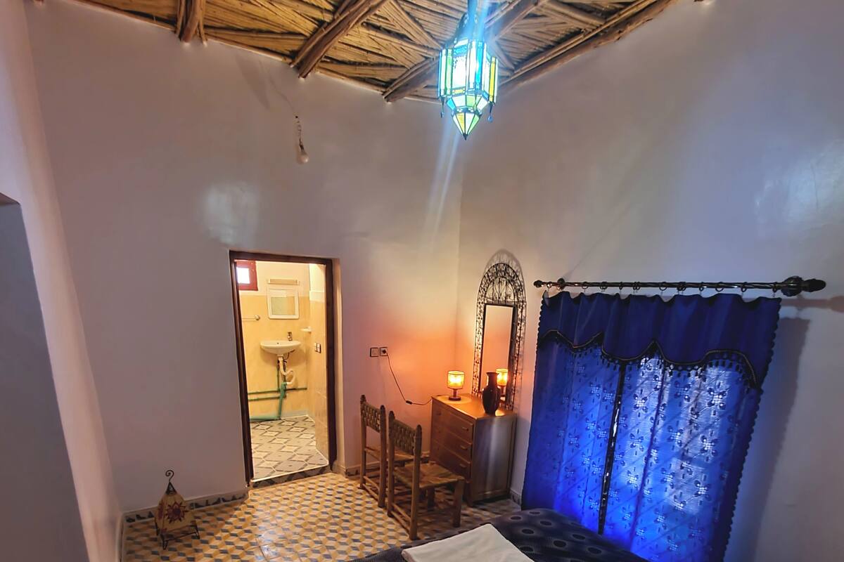 Ein Schlafzimmer mit Schachbrettboden, einem Bett mit gemustertem Überwurf, einem Nachttisch mit Lampe, einem Spiegel und einem Fenster mit blauen Vorhängen.