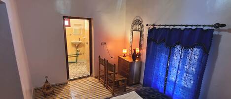A bedroom with a checkered floor, a bed with a patterned bedspread, a nightstand with a lamp, a mirror, and a window with blue curtains.