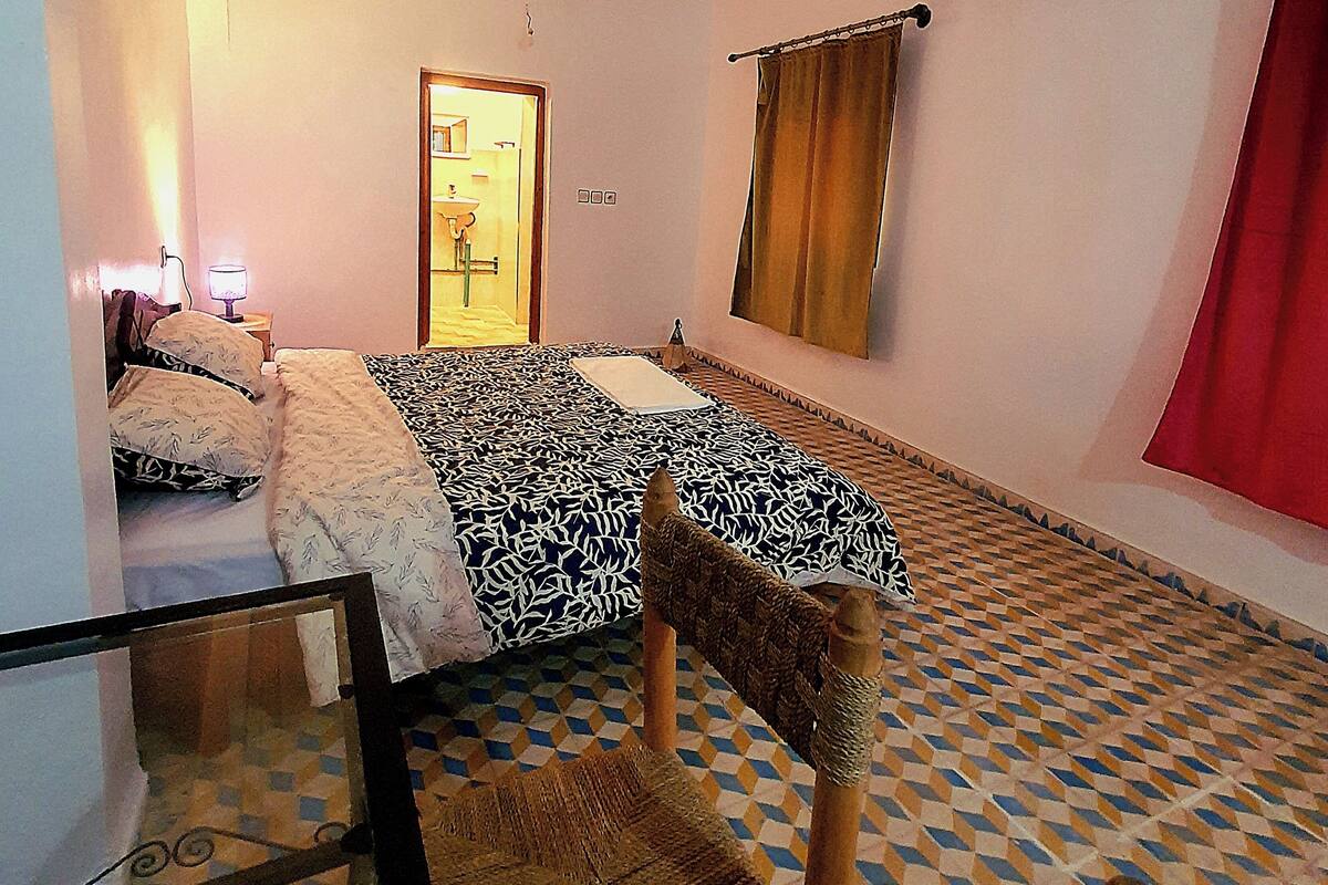 Ein Zimmer mit einem Bett, einem Sessel, einem Tisch, einem Fenster mit Vorhängen und einer Tür zu einem Badezimmer.