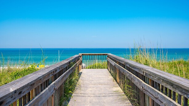 Ein hölzerner Steg führt zu einem Strand mit klarem, blauem Wasser und blauem Himmel.
