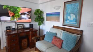 Una sala de estar con un escritorio de madera, un sofá con cojines estampados, un televisor y cuadros en las paredes.