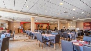 Ein geräumiges Restaurant mit mehreren Tischen und Stühlen, einem Küchenbereich im Hintergrund und einem gefliesten Boden.