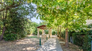 Ein gepflasterter Weg führt zu einer Laube in einer parkähnlichen Umgebung mit Bäumen und Grün.