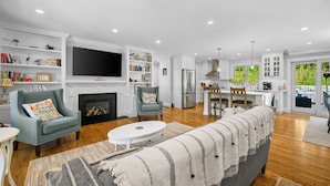 A spacious living room with a fireplace, a television, and a kitchen area with a dining table. - Coastal Escape: Steps to Red River Beach (Harwich Port)