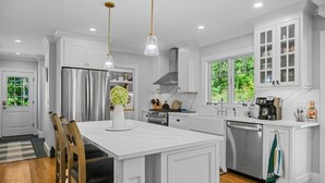 A modern kitchen with white cabinetry, a central island, stainless steel appliances, and pendant lighting. - Coastal Escape: Steps to Red River Beach (Harwich Port)