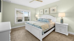 Une chambre à coucher comprenant un lit, deux tables de chevet, un ventilateur de plafond, une fenêtre avec des stores et des œuvres d’art murales.