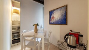 A dining area with a white table and chairs, a vase with flowers, a painting on the wall, and kitchen appliances on a rack.