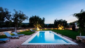 A rectangular swimming pool with a brick border, surrounded by a grassy area with trees and a stone wall.