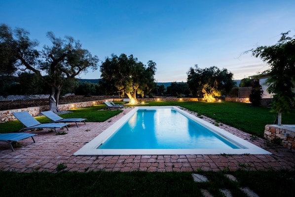 A rectangular swimming pool with a brick border, surrounded by a grassy area with trees and a stone wall.