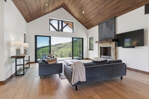 A spacious living room with a large gray sofa, a coffee table, and a fireplace. There is a large window offering a view of a green landscape.