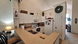 A kitchen with white cabinets, a granite countertop, a stove, a microwave, and a refrigerator. There is a sink, a chair, and a table with a lamp.