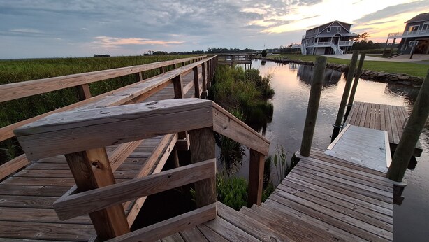 A wooden boardwalk leading to a dock over a calm water body, with houses in the background and a cloudy sky.