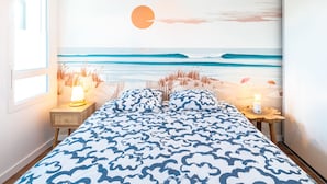 A bedroom with a beach mural, a bed with a blue and white patterned bedspread, bedside tables with lamps, and a window with blinds.