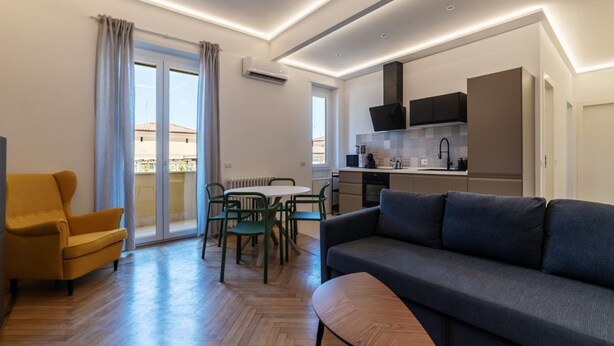 A modern living room with a sofa, a dining table with chairs, and a kitchen area with cabinets and a stove. - GuestHost - Comfortable apartment located on the seventh floor, of nine in total, equipped with a lift (NOT suitable for disabled people). There are steps at the entrance.In 65 m2 of space it accommodates 2 people with a double bedroom.The location is ex (Roma)