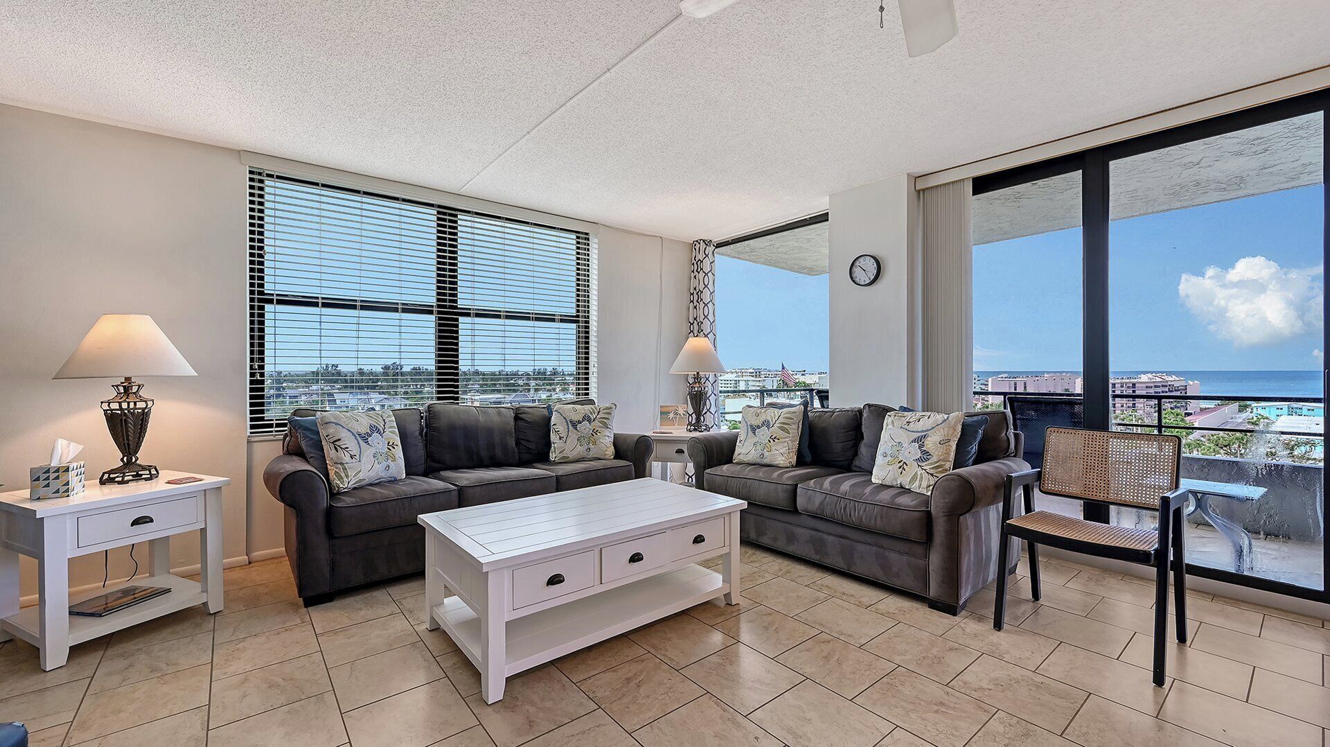 A spacious living room with two sofas, a coffee table, and a side table with lamps. Large windows offer a view of the city and ocean.