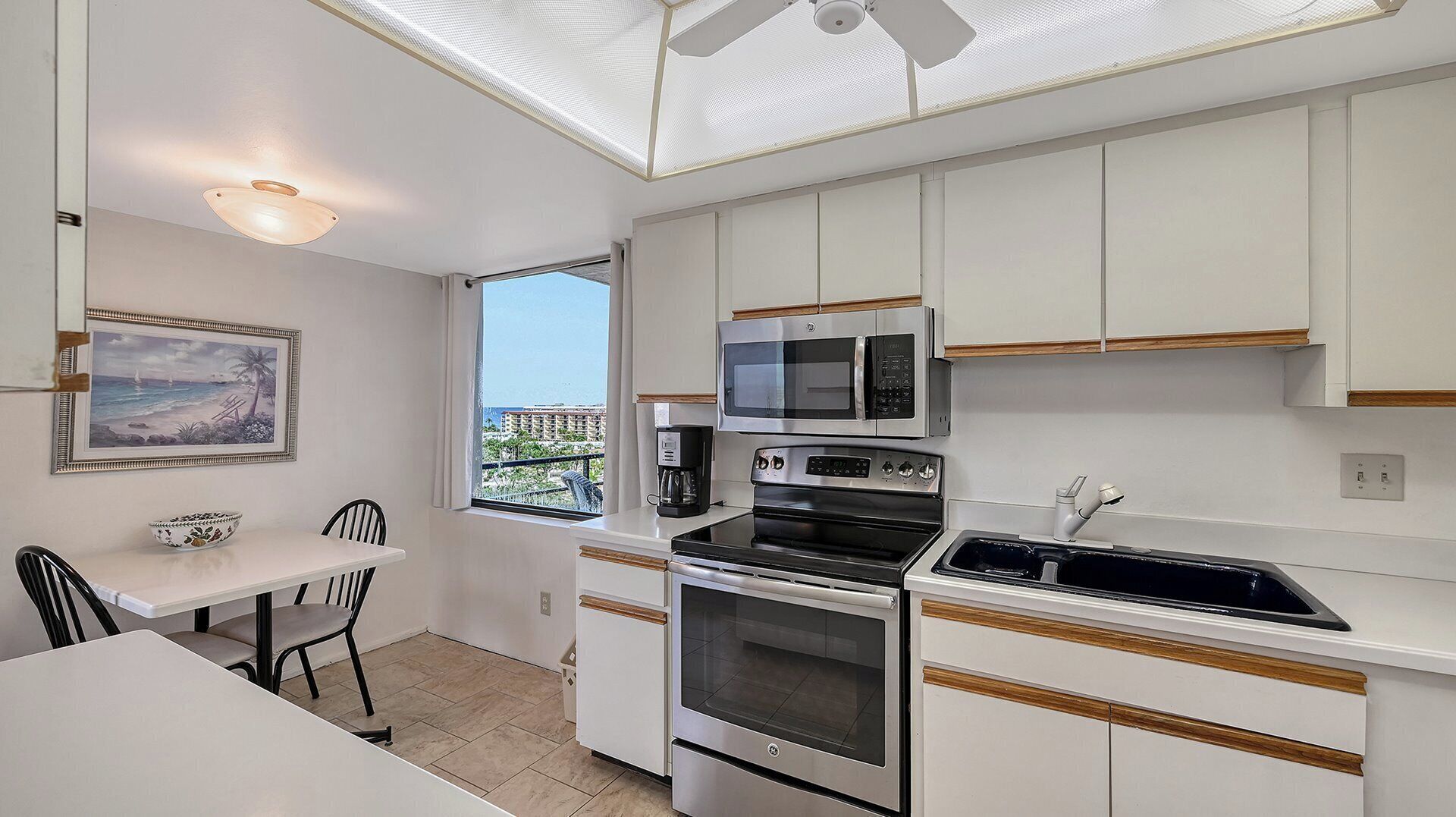 A compact kitchen with white cabinets, a built-in oven, a microwave, and a coffee maker. There's a dining area with a table and chairs, and a window offering a view of buildings.