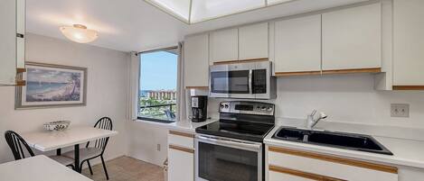 A compact kitchen with white cabinets, a built-in oven, a microwave, and a coffee maker. There's a dining area with a table and chairs, and a window offering a view of buildings.