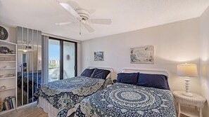 A bedroom with two beds, a ceiling fan, a nightstand with a lamp, a bookshelf, and a view of the outside. - Anchorage 804 in Siesta Key, FL (Siesta Key)