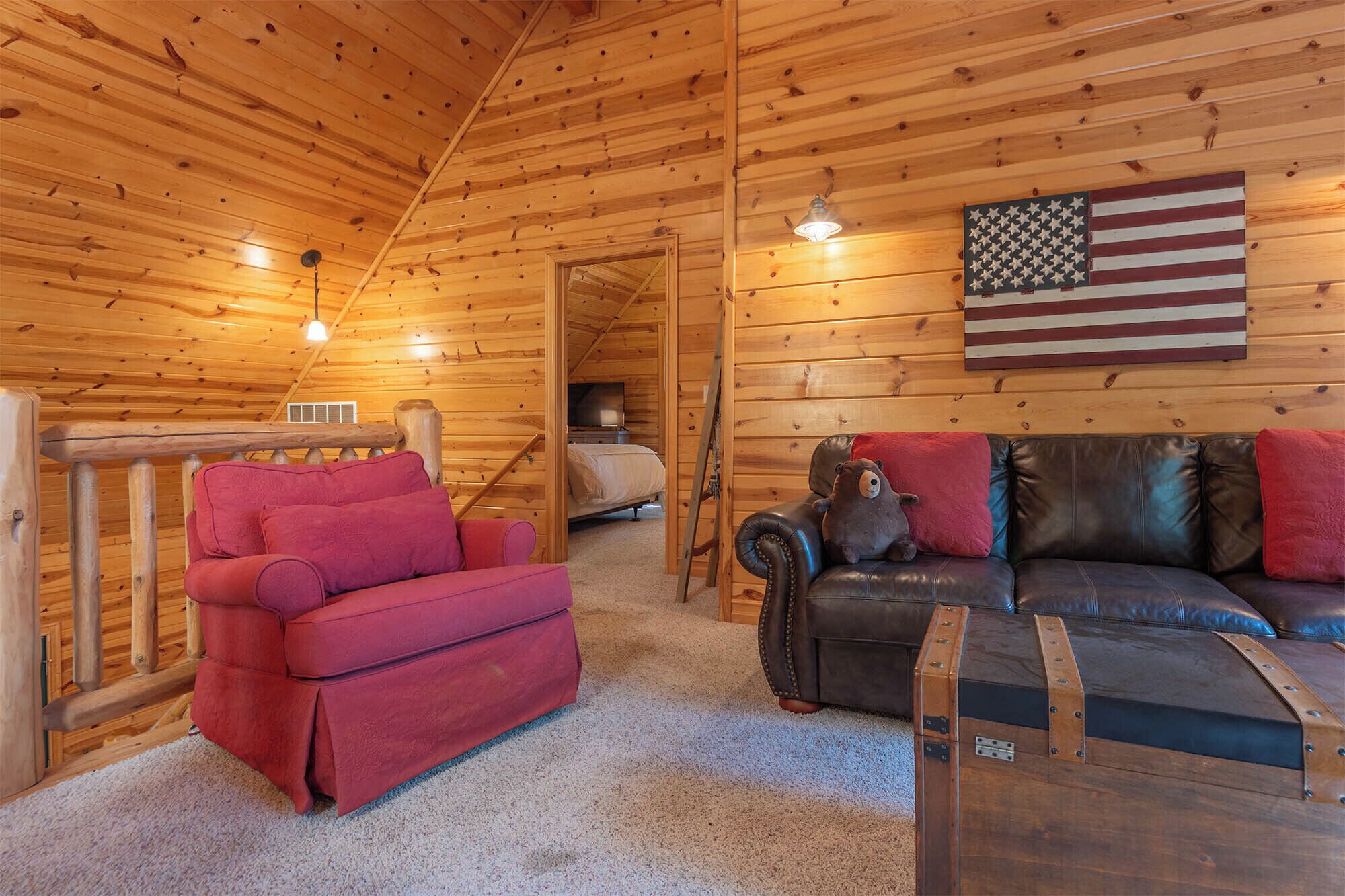 A cozy cabin interior with a wooden ceiling, a red sofa, a black leather couch, and a large American flag on the wall.