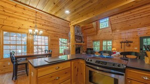 A wooden cabin interior with a kitchen featuring a large stove, wooden cabinets, and a dining area with chairs.