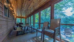 A covered deck with wooden flooring, a table, chairs, and large windows offering a view of the surrounding forest.