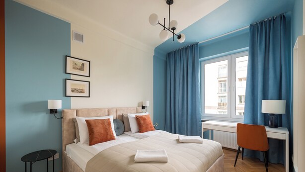 A bedroom with a large bed, a desk, and a chair. There are two framed pictures on the wall and a window with curtains. - Warsaw Dream Apartment (Warszawa)