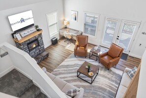 A spacious living room with a fireplace, a desk, leather chairs, and a large TV displaying a logo.