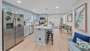 Una cocina moderna con electrodomésticos de acero inoxidable, una isla central con taburetes, armarios azul claro y una zona de comedor con mesa y sillas.