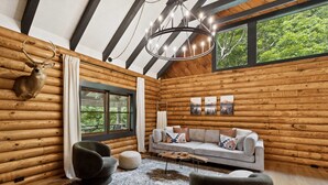 Ein Holzblockhaus-Interieur mit einer Couch, Sesseln, einer Chandelieber und einem an der Wand befestigten Hirschkopf.