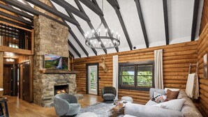 Ein Blockhaushaus-Interieur mit Kamin aus Naturstein, Holzbalken, einer Kristallleuchtre und einer gemütlichen Sitzecke.
