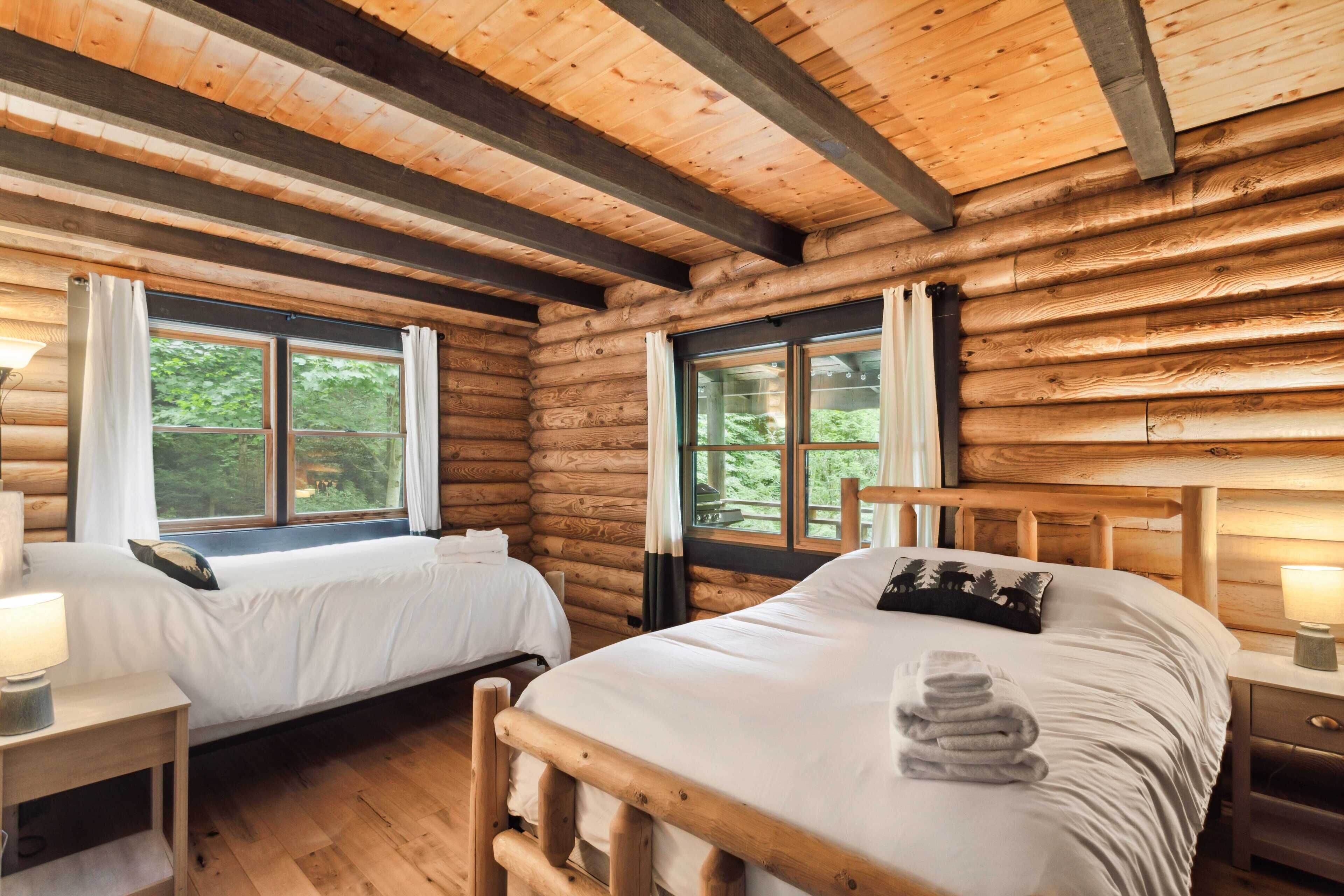 Une chambre de style rustique avec deux lits, des poutres apparentes, de grandes fenêtres et une tête de lit en bois.
