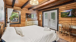 Ein Schlafzimmer in einer Blockhütte mit einem großen Bett, Holzbalken an der Decke, einem Schreibtisch und einem Fenster mit Blick ins Grüne.