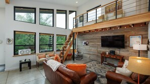 A spacious living area with a high ceiling, large windows, a wooden staircase, a leather sofa, a TV, and a fireplace. - Gallatin River Retreat: 3BR Home with Hot Tub, Fly Fishing & Stunning Views (Gallatin Gateway)