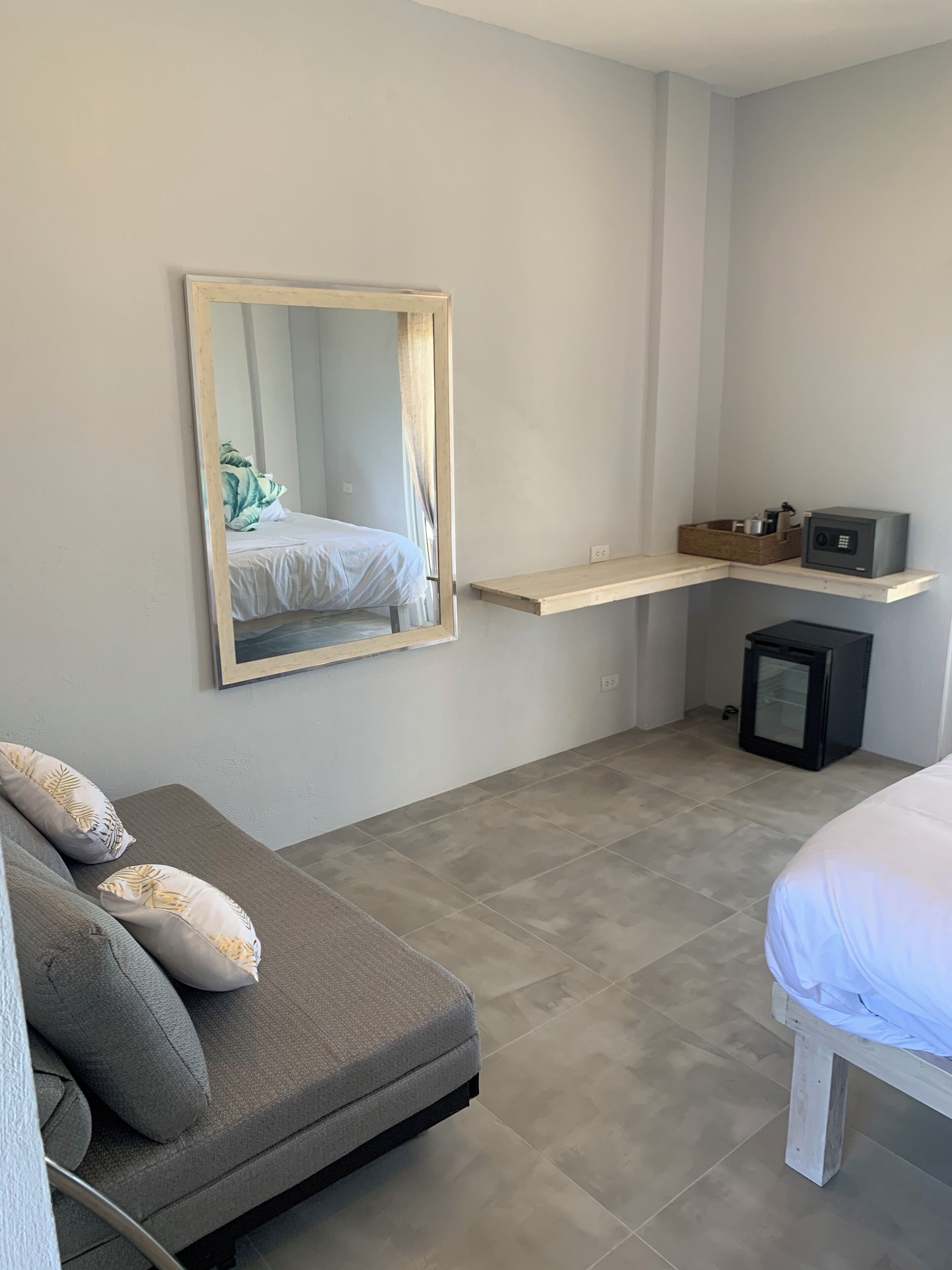 A modern bedroom with a grey sofa, a bed with white bedding, a large mirror, a small black safe, and a wooden shelf with a basket.