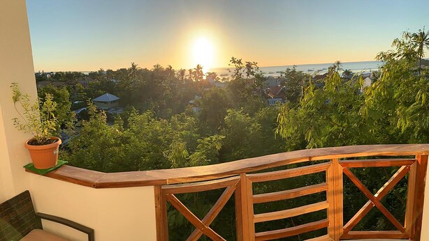 A balcony with a wooden railing overlooking a residential area and a body of water during sunset. - Crown Malapascua (Daanbantayan)