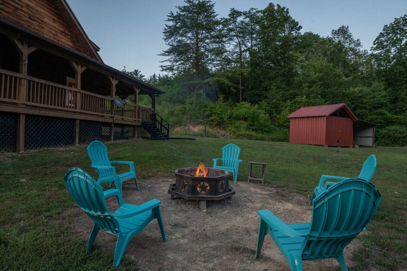 A deck with outdoor seating and a fire pit in a grassy area with trees and a red barn in the background.