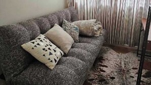 A living room with a dark grey sofa, patterned cushions, a patterned rug, and a curtain with vertical stripes. - Westgate Three and one small room house 🏠  (Auckland)