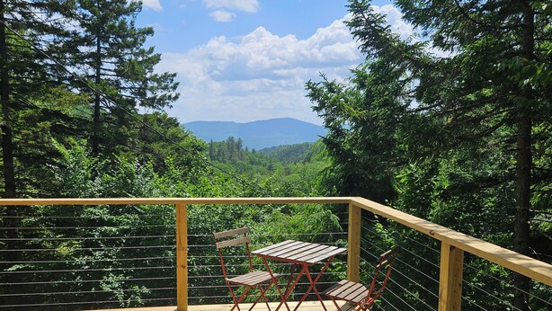 Une terrasse en bois, agrémentée d’une table et de chaises, entourée d’une rambarde en métal, et offrant une vue sur un paysage boisé avec des montagnes à l’horizon.