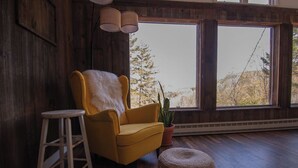 Une pièce avec un fauteuil jaune, un plaid blanc et moelleux, une plante en pot, un tabouret en bois et un pouf tressé.