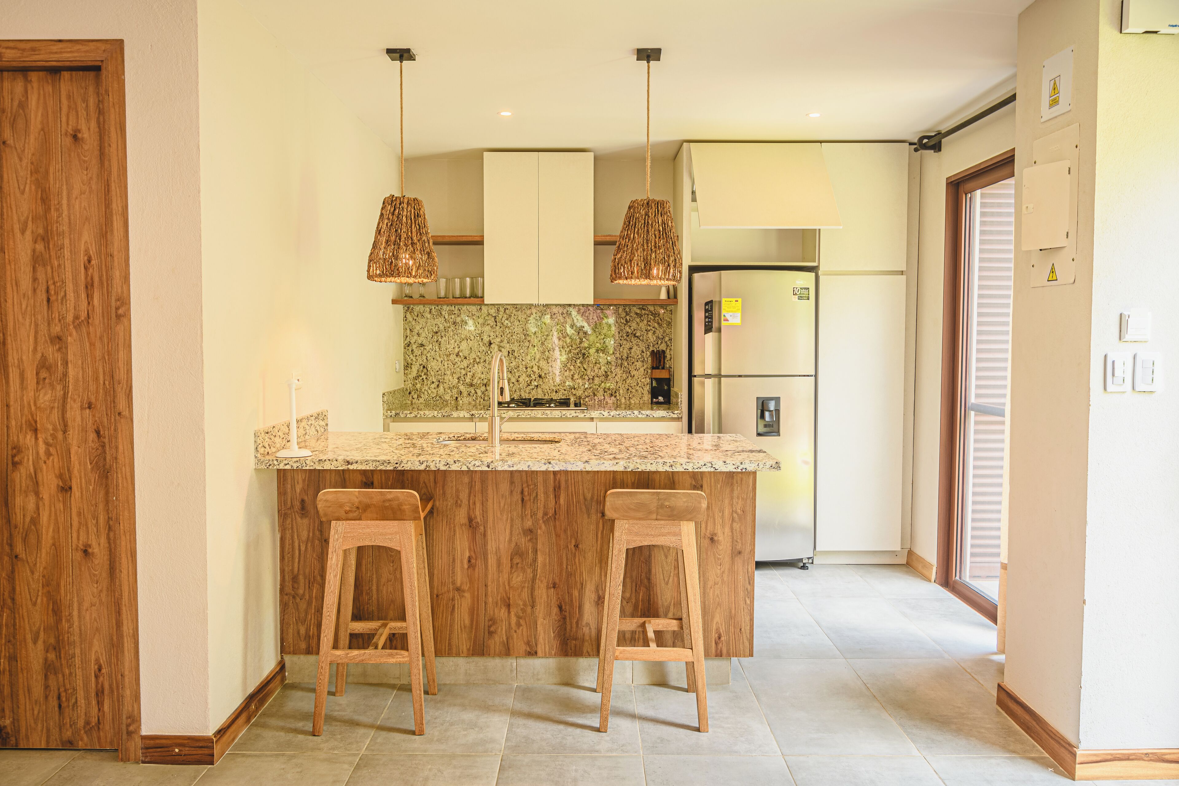 A kitchen with a central island, two bar stools, pendant lights, a refrigerator, and a tiled backsplash.