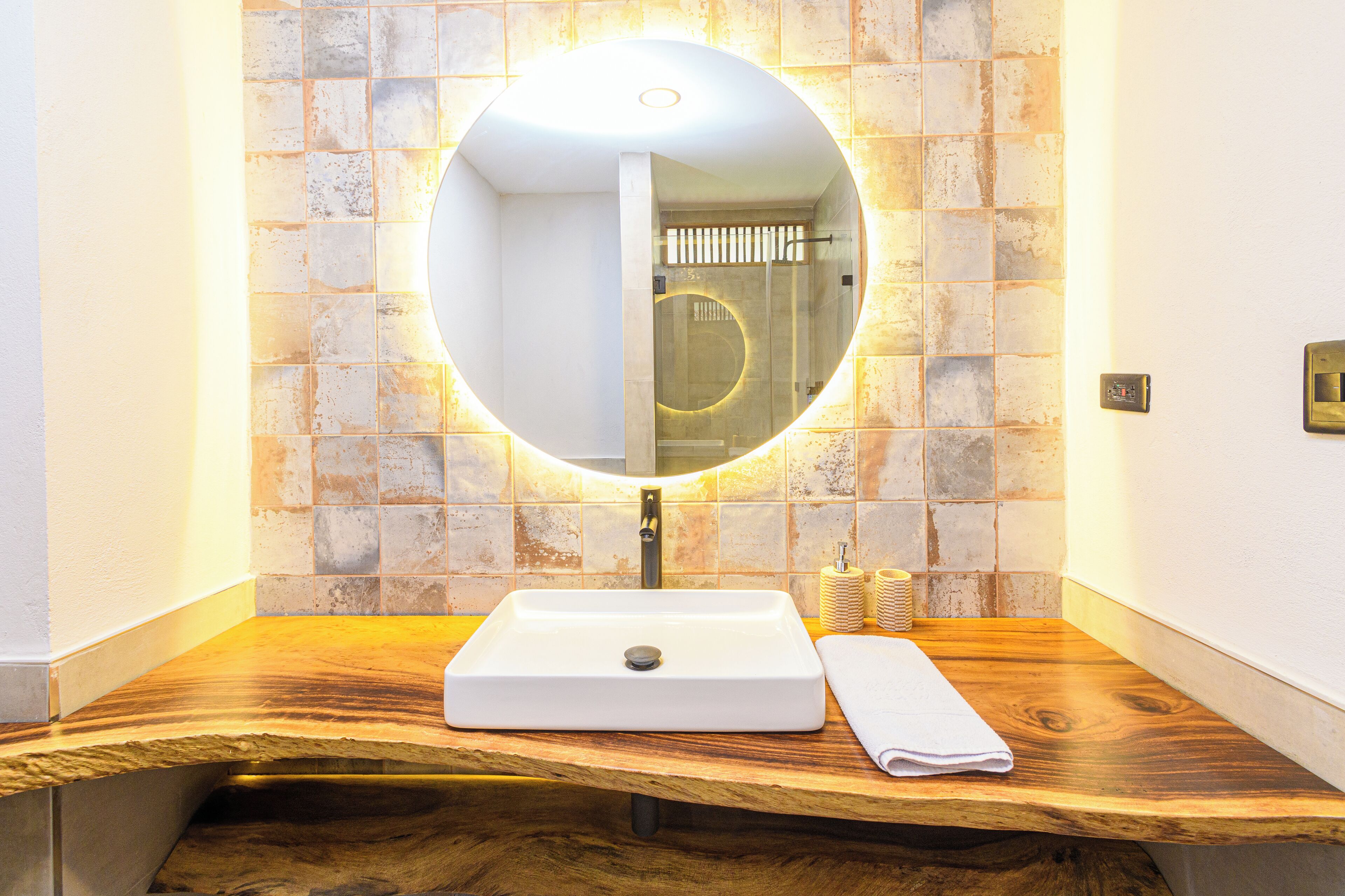 Un baño con un tocador de madera, un lavabo blanco, un espejo redondo con luz y una pared con azulejos.