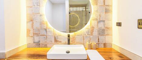 Un baño con un tocador de madera, un lavabo blanco, un espejo redondo con luz y una pared con azulejos.