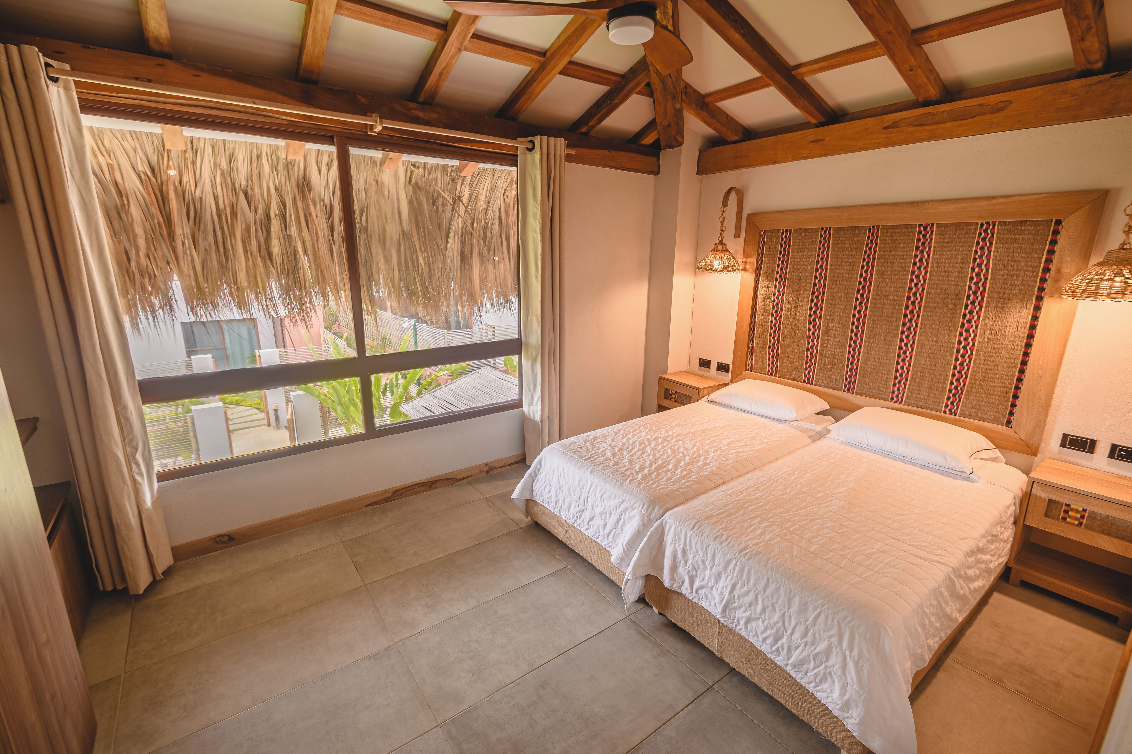 A bedroom with a large bed, wooden ceiling beams, a window with curtains, and a bedside lamp.