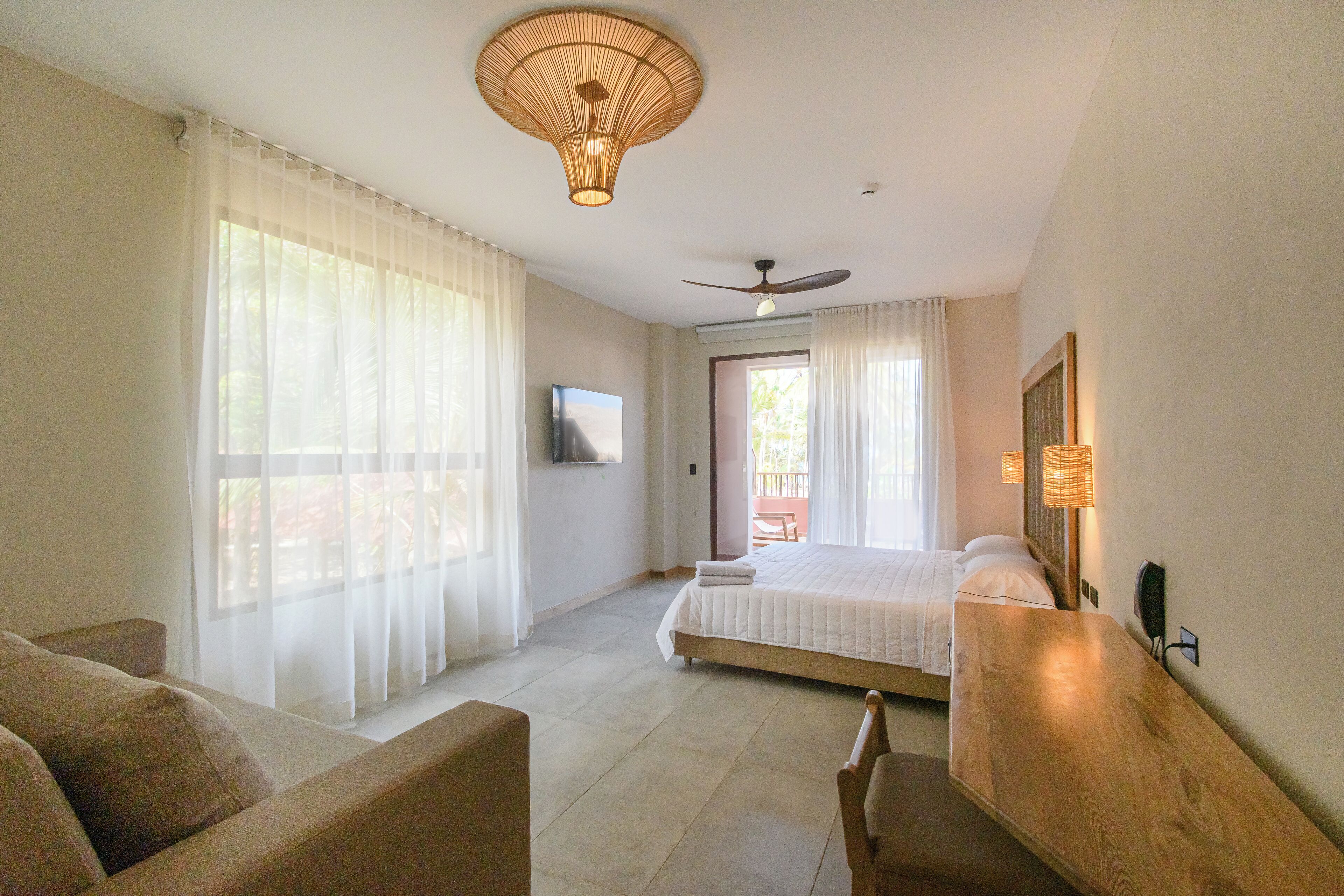 Un dormitorio con cama, ventilador de techo, televisor y ventana con cortinas traslúcidas.