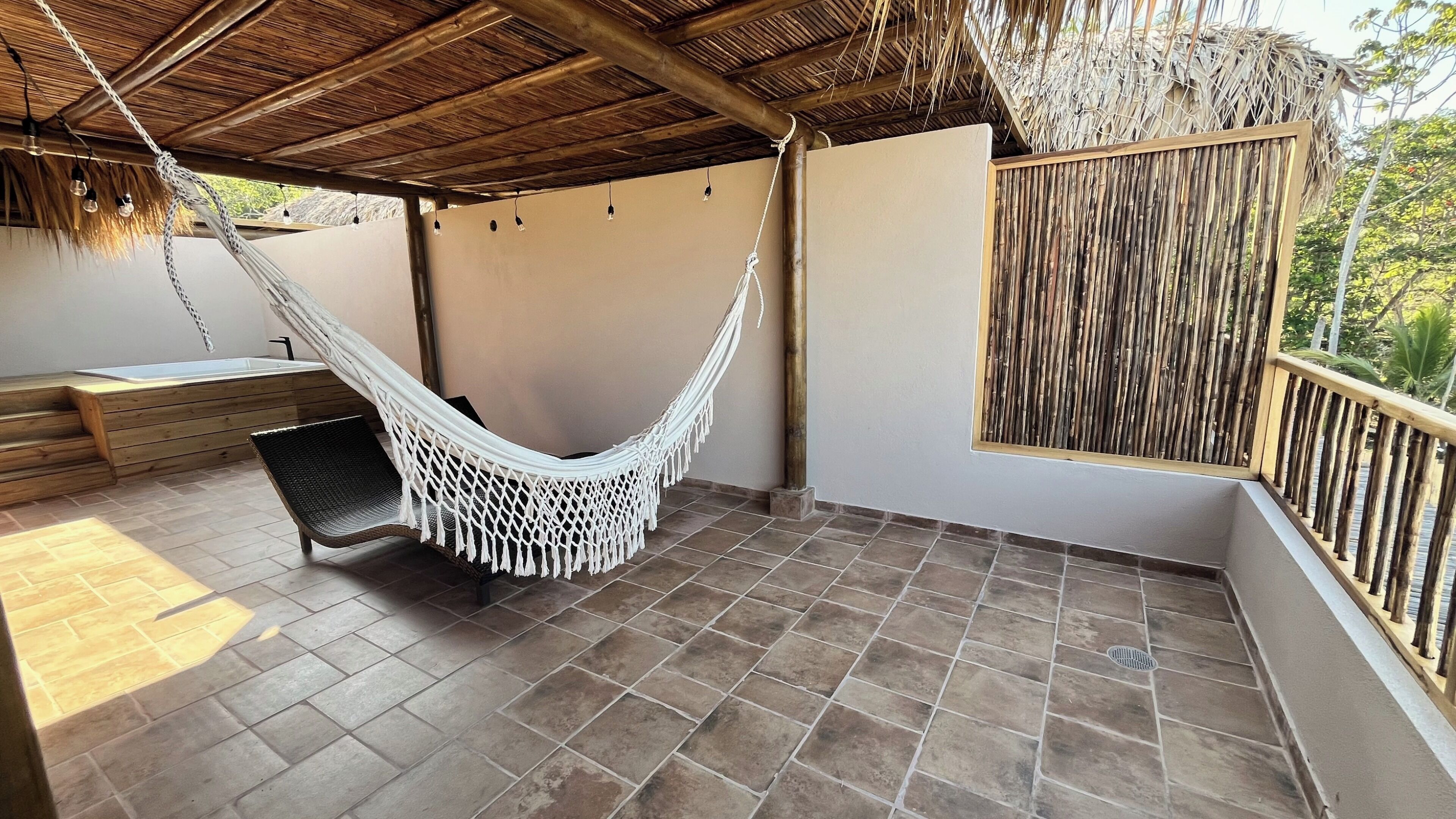A hammock under a thatched roof, a wooden bench, and a view of greenery.