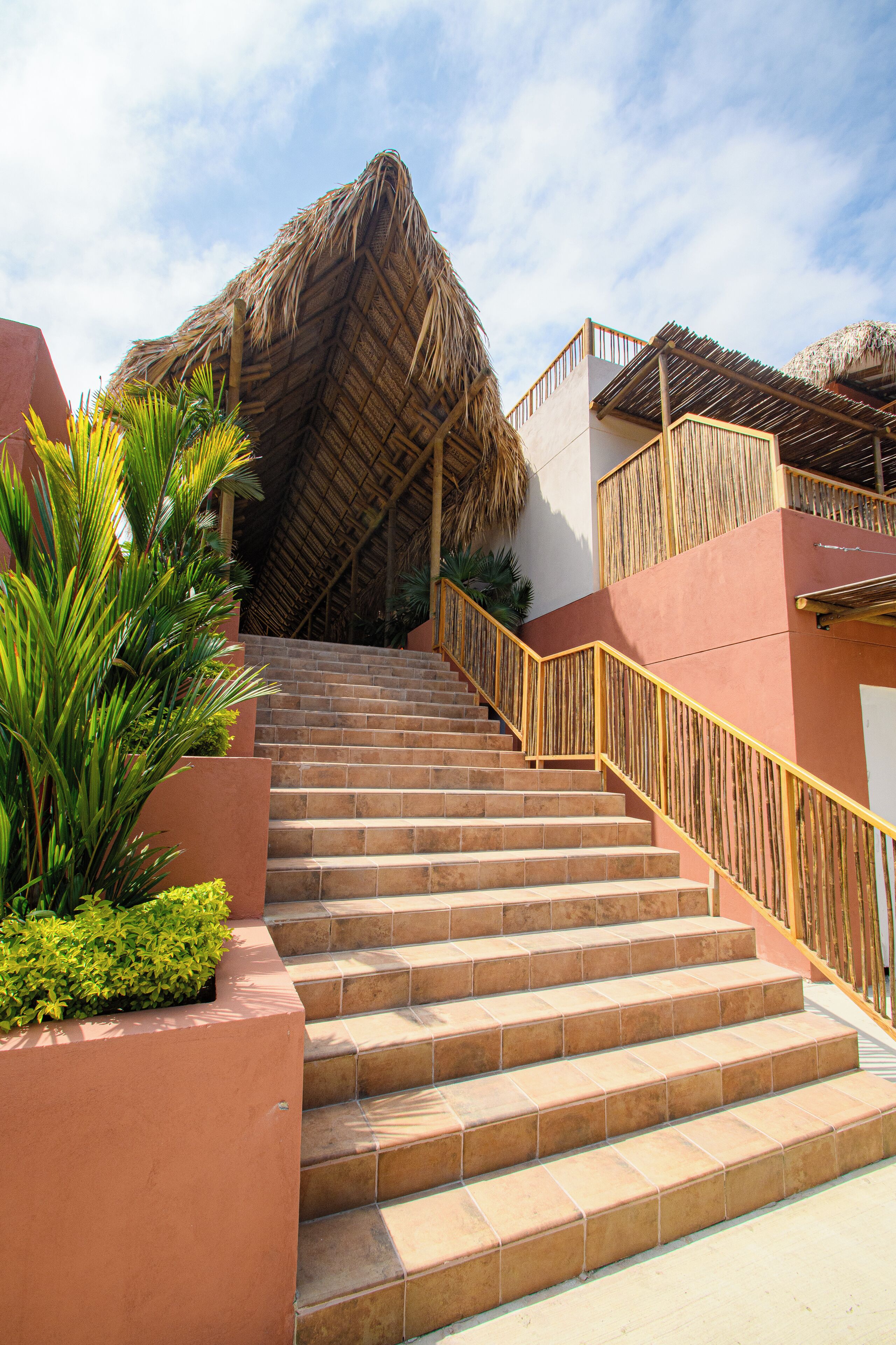 A staircase leading to a building with a thatched roof, surrounded by greenery and a clear sky.